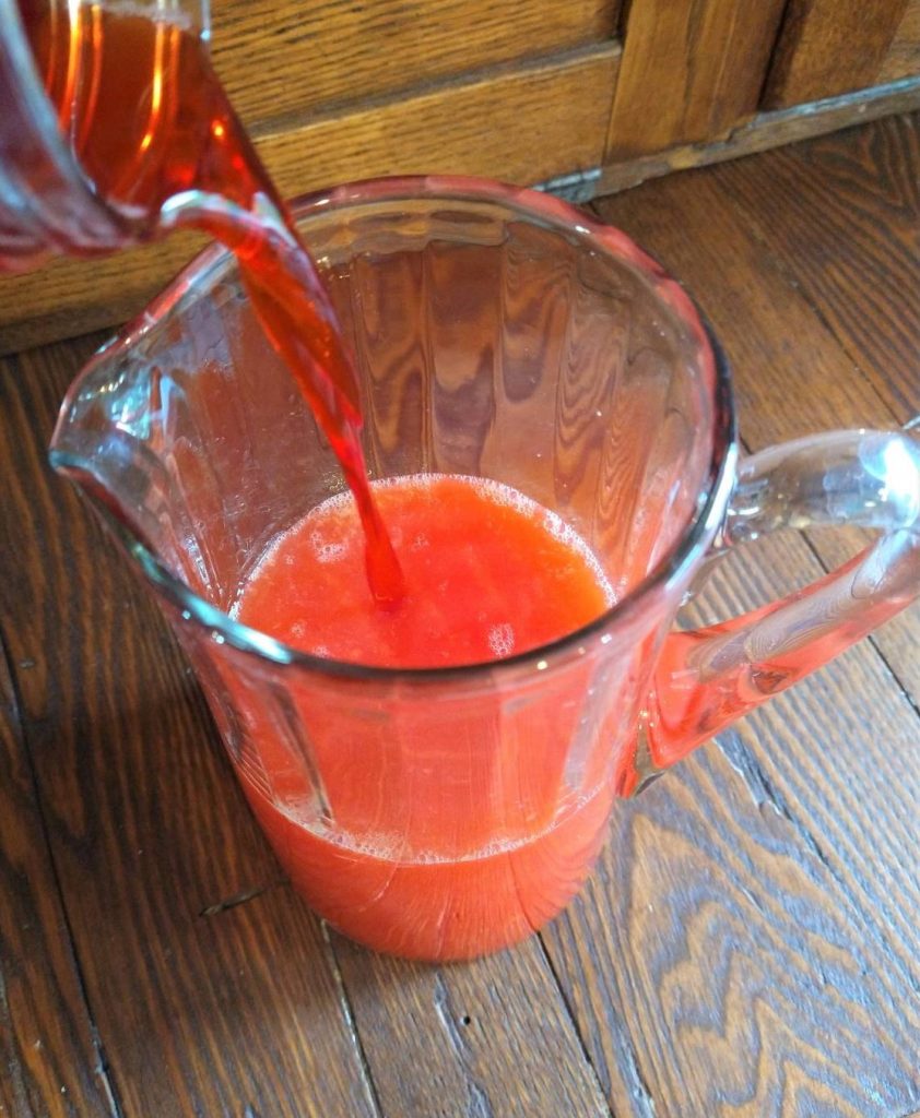 strawberry syrup pouring into a glass pitcher of juice 