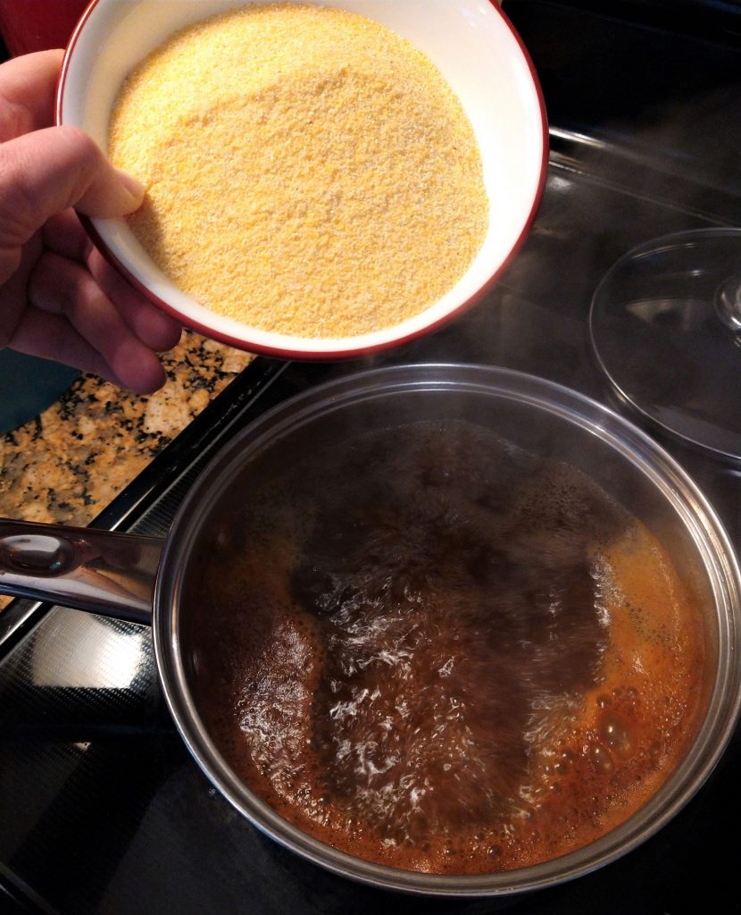 hand holding a bowl of cornmeal over a pot of boiling water spiced with chili powder