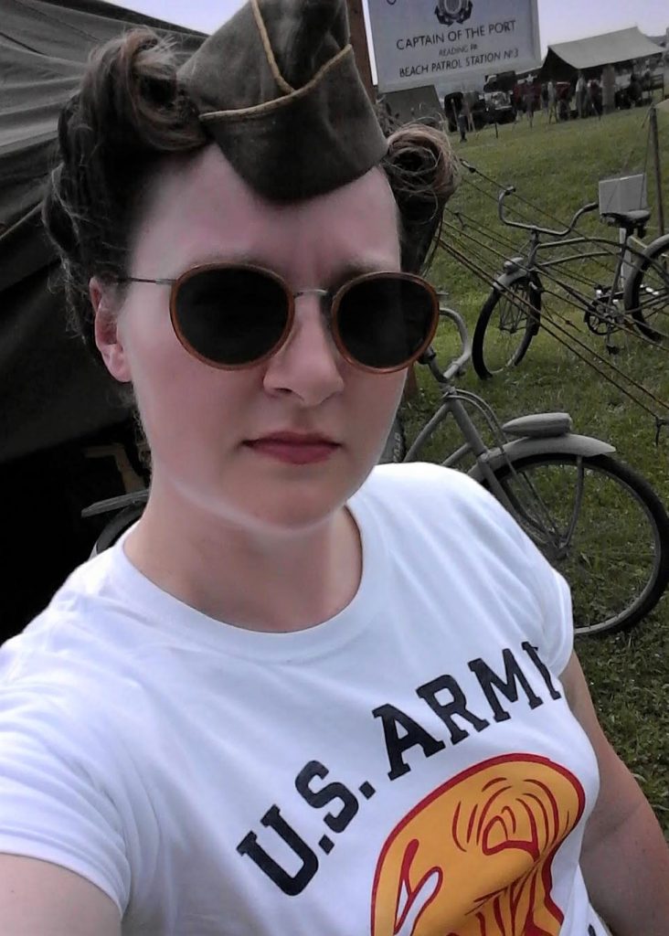 Sarah Lee wearing a WWII WAC t-shirt, cap and vintage=-tyle shades with WWII tents and bicycles in the background