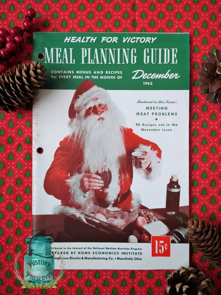 an old copy of Health for Victory Meal Planning Guide dated December 1943 with Santa Claus on the cover sits on a red Christmas fabric studded with green Christmas trees. Pinecones and red berries decorate the corners of the frame.