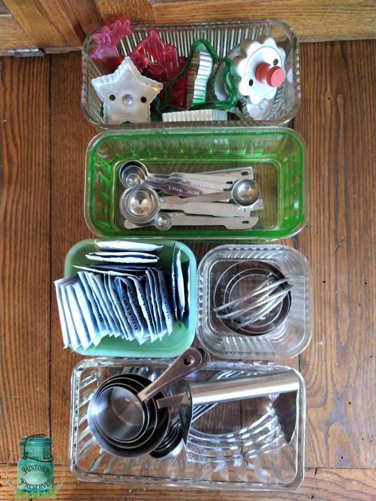 old glass dishes on a wooden table holding various things like measuring spoons and cups, biscuit cutters, tea bags and cookie cutters