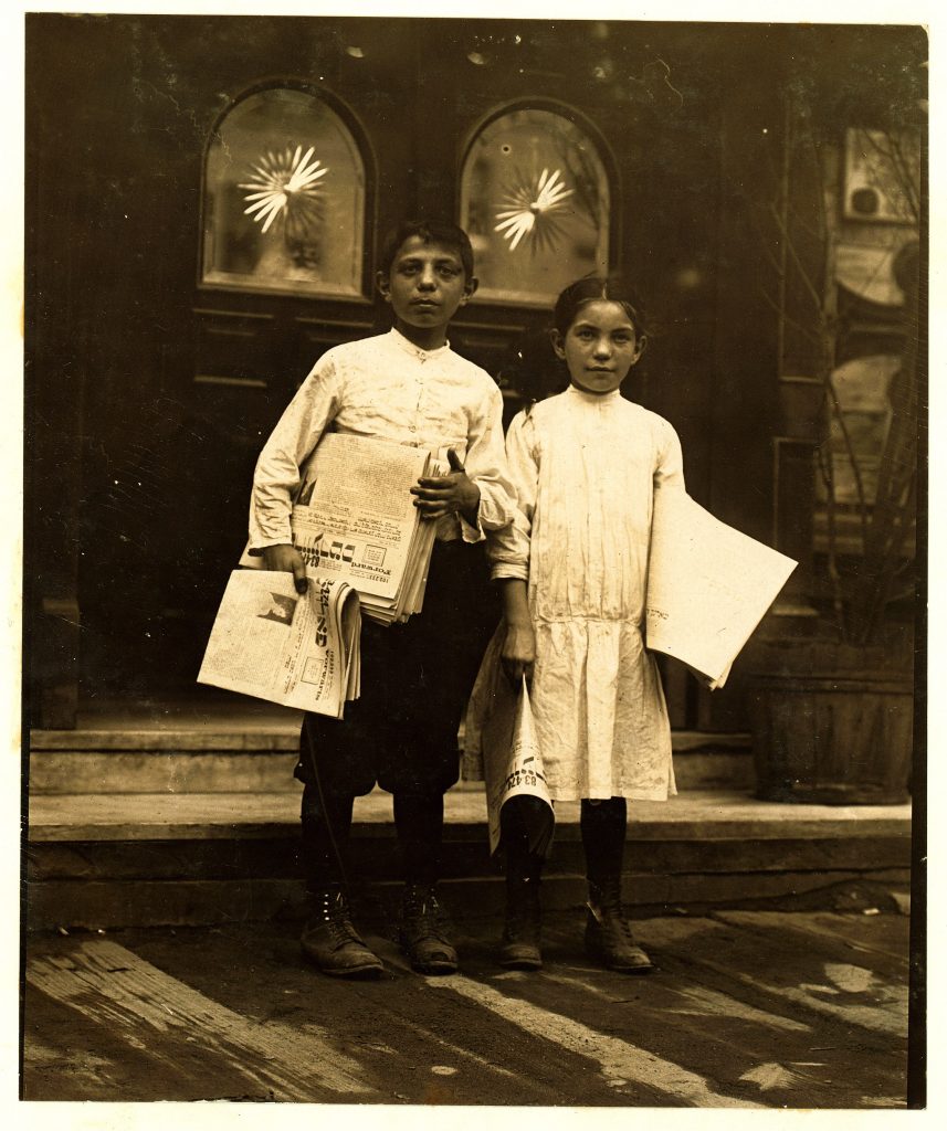 an old photograph from 1910 shows a boy and girl dressed in old-fashioned clothes looking at the camera and holding stacks of newspapers. Lewis Hine is the photographer.